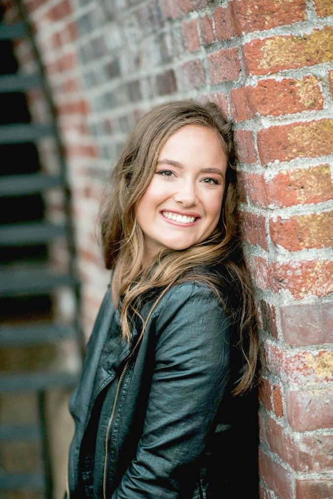 young brunette woman standing against brick wall in black leather jacket smiling at camera