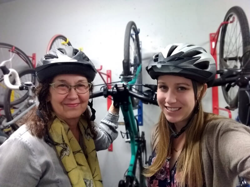 two women, one younger and one middle-aged, with helpmets on standing in front of hanging bikes smiling for camera