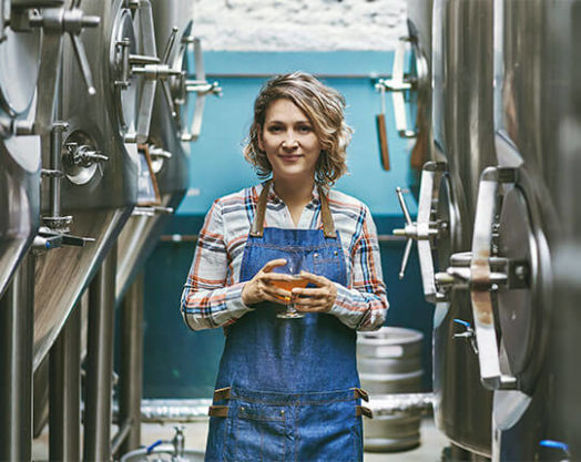 Sonriente mujer cervecera artesana de pie con un vaso de cerveza extraído directamente de una cuba para su degustación.