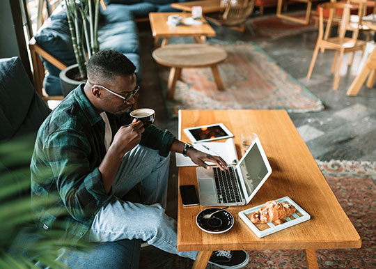 Side view of a young man sitting in a cafe, casually working using his laptop and drinking a cup of coffee.